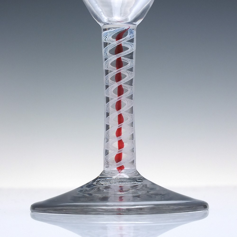 An eighteenth-century clear lead glass ale flute featuring a flared rim, floral hop wheel engravings, and a central stem containing a red and opaque white spiral twist. View III