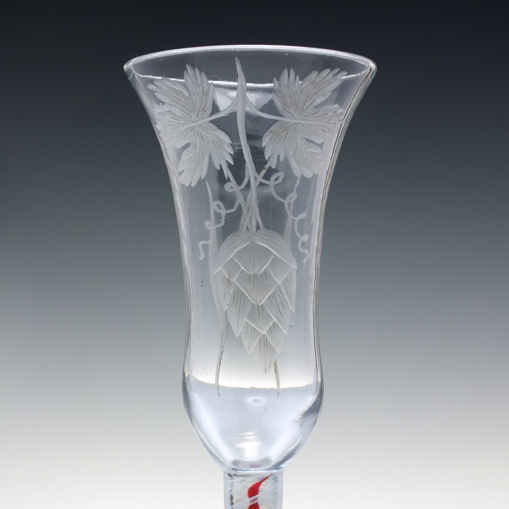 An eighteenth-century clear lead glass ale flute featuring a flared rim, floral hop wheel engravings, and a central stem containing a red and opaque white spiral twist. View IV