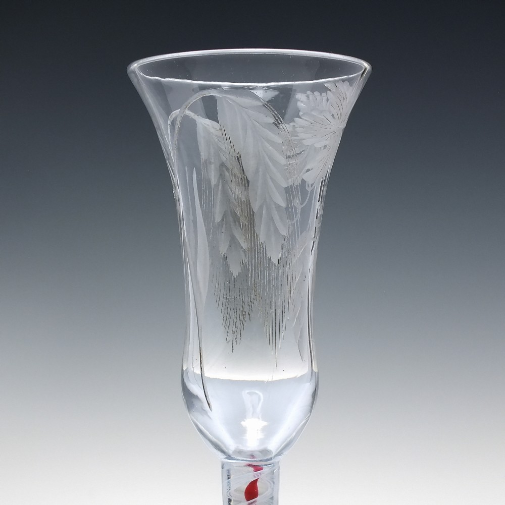 An eighteenth-century clear lead glass ale flute featuring a flared rim, floral hop wheel engravings, and a central stem containing a red and opaque white spiral twist. View V
