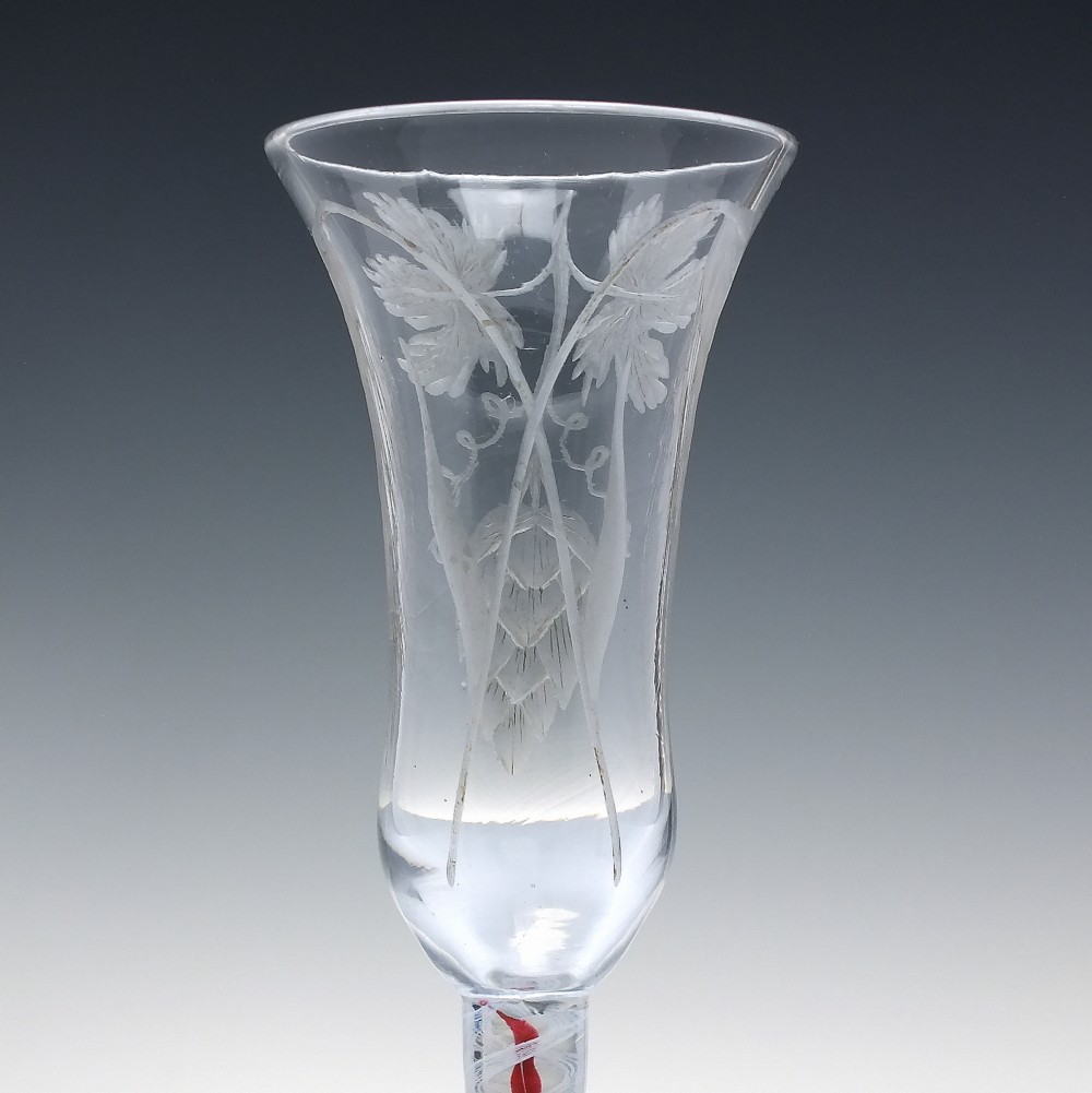 An eighteenth-century clear lead glass ale flute featuring a flared rim, floral hop wheel engravings, and a central stem containing a red and opaque white spiral twist. View VI