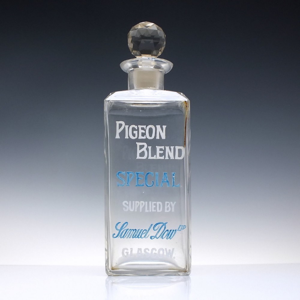 A rectangular clear glass whisky decanter featuring white and blue enamelled text reading Pigeon Blend Special and topped with a faceted glass stopper.