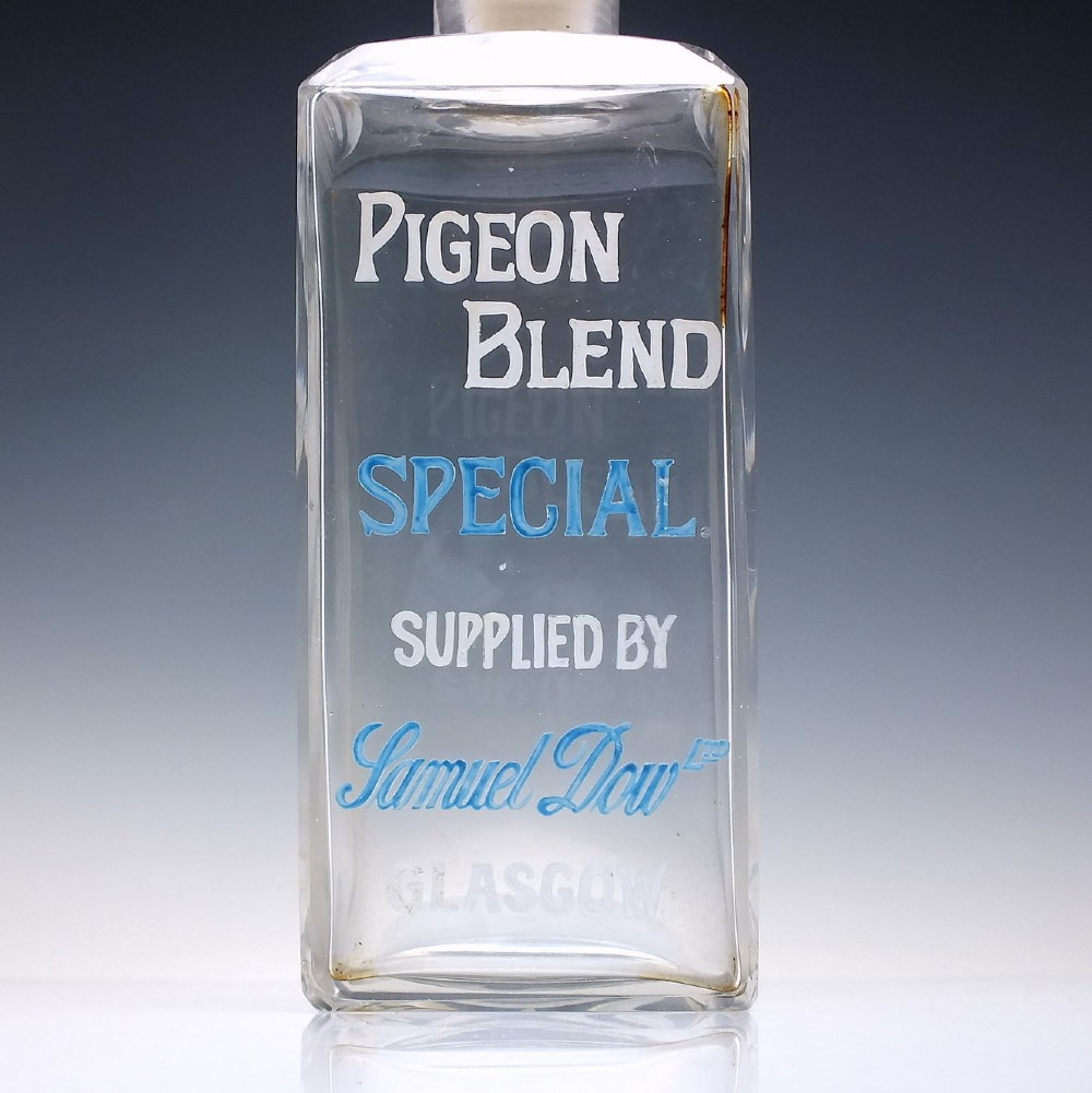 A rectangular clear glass whisky decanter featuring white and blue enamelled text reading Pigeon Blend Special and topped with a faceted glass stopper. View IV