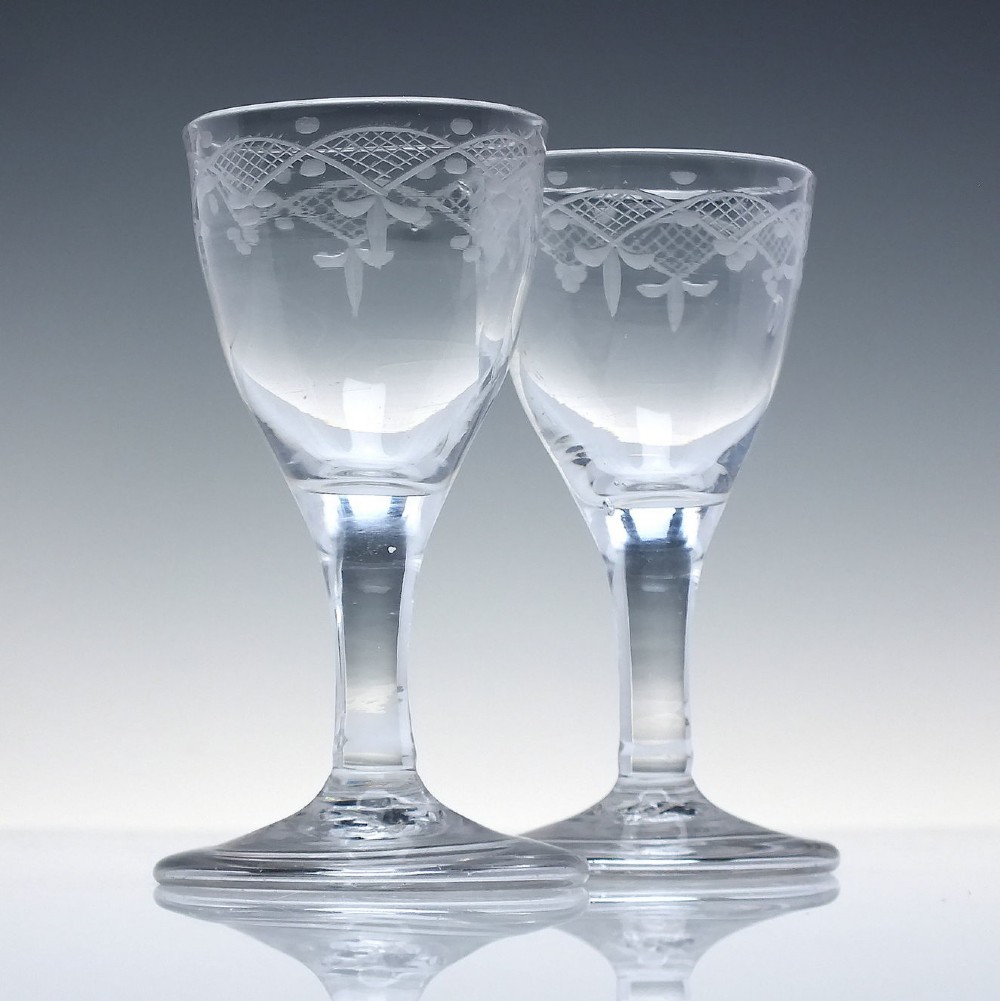 A pair of 18th-century clear glass drams featuring wheel-engraved draped swag borders, plain stems, and wide folded feet.