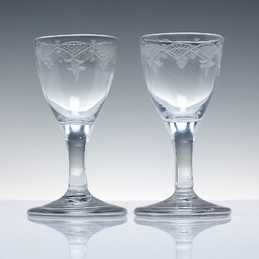 A pair of 18th-century clear glass drams featuring wheel-engraved draped swag borders, plain stems, and wide folded feet. View II