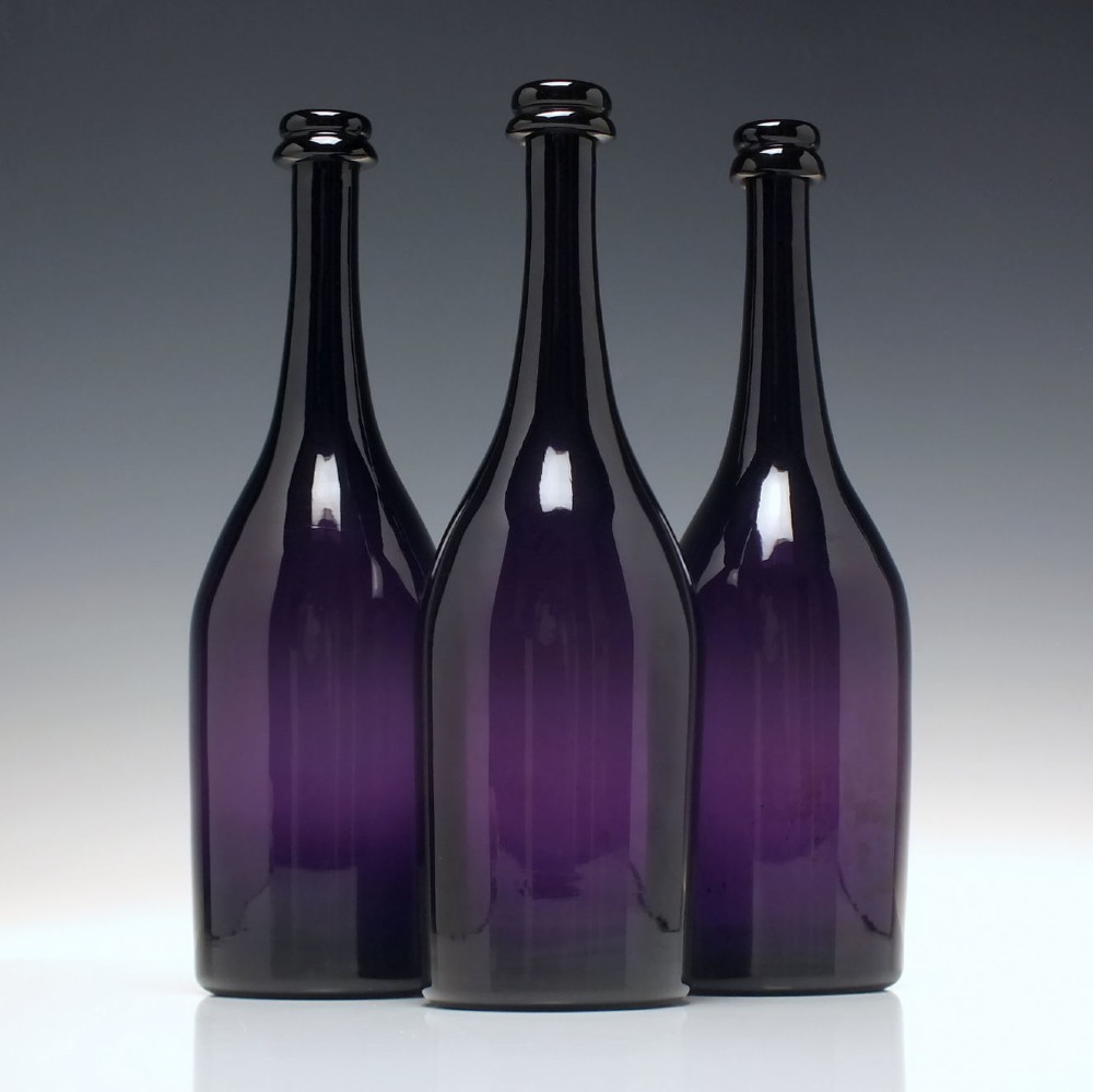 A set of three Victorian hand-blown deep purple amethyst glass wine serving bottles featuring long tapered necks and double ring lip collars.