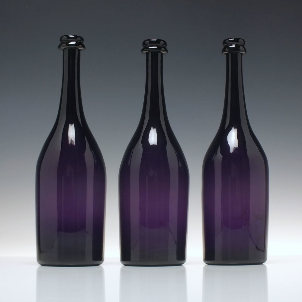 A set of three Victorian hand-blown deep purple amethyst glass wine serving bottles featuring long tapered necks and double ring lip collars. View II