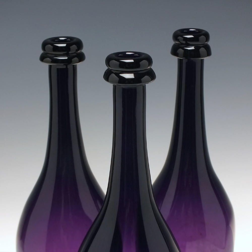 A set of three Victorian hand-blown deep purple amethyst glass wine serving bottles featuring long tapered necks and double ring lip collars. View III