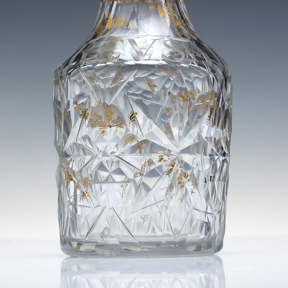 A clear cut glass decanter with gilded lettering on the shoulder, a diamond facetted body, and a facetted glass stopper. View VI