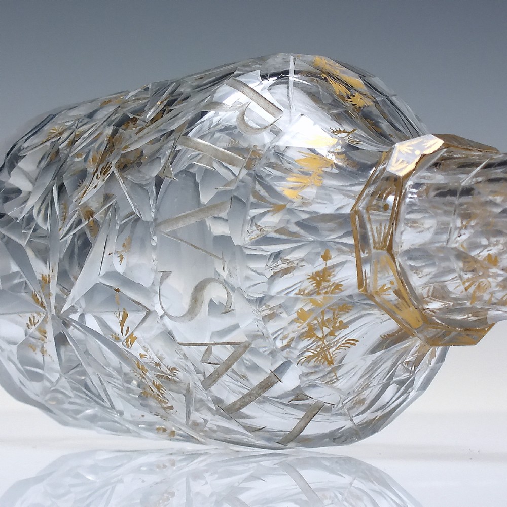 A clear cut glass decanter with gilded lettering on the shoulder, a diamond facetted body, and a facetted glass stopper. View IX