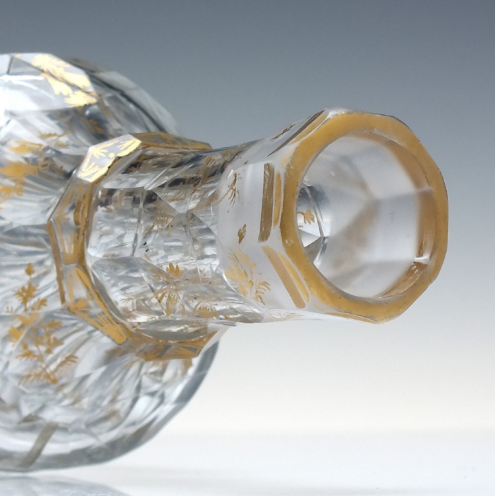 A clear cut glass decanter with gilded lettering on the shoulder, a diamond facetted body, and a facetted glass stopper. View X