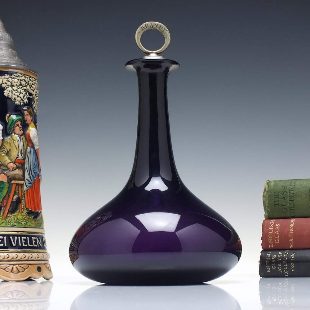 A deep purple amethyst glass decanter featuring a wide bulbous base, elongated neck, and a metal ring stopper engraved with the word BRANDY.
