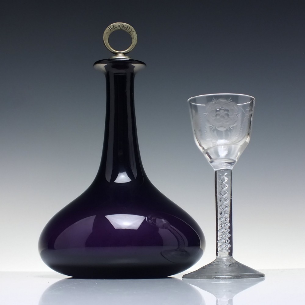 A deep purple amethyst glass decanter featuring a wide bulbous base, elongated neck, and a metal ring stopper engraved with the word BRANDY. View II