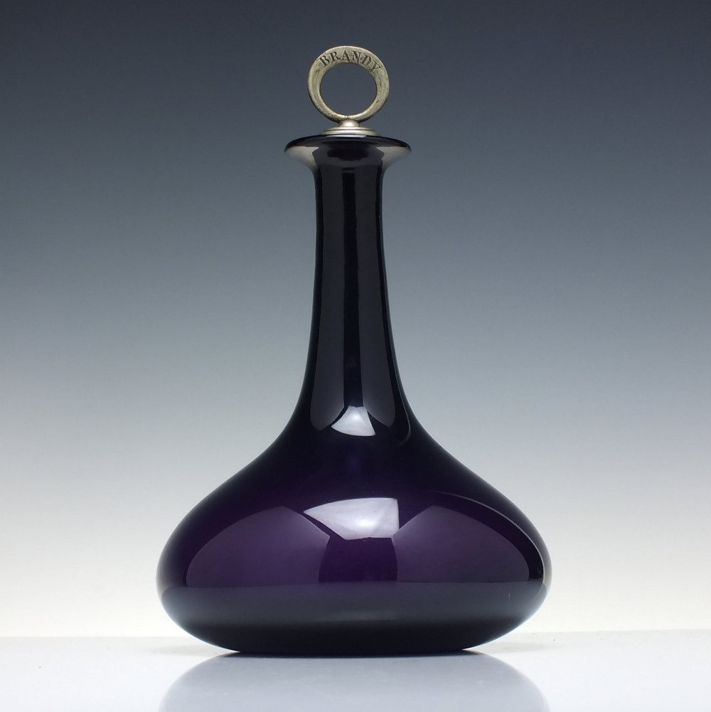 A deep purple amethyst glass decanter featuring a wide bulbous base, elongated neck, and a metal ring stopper engraved with the word BRANDY. View III