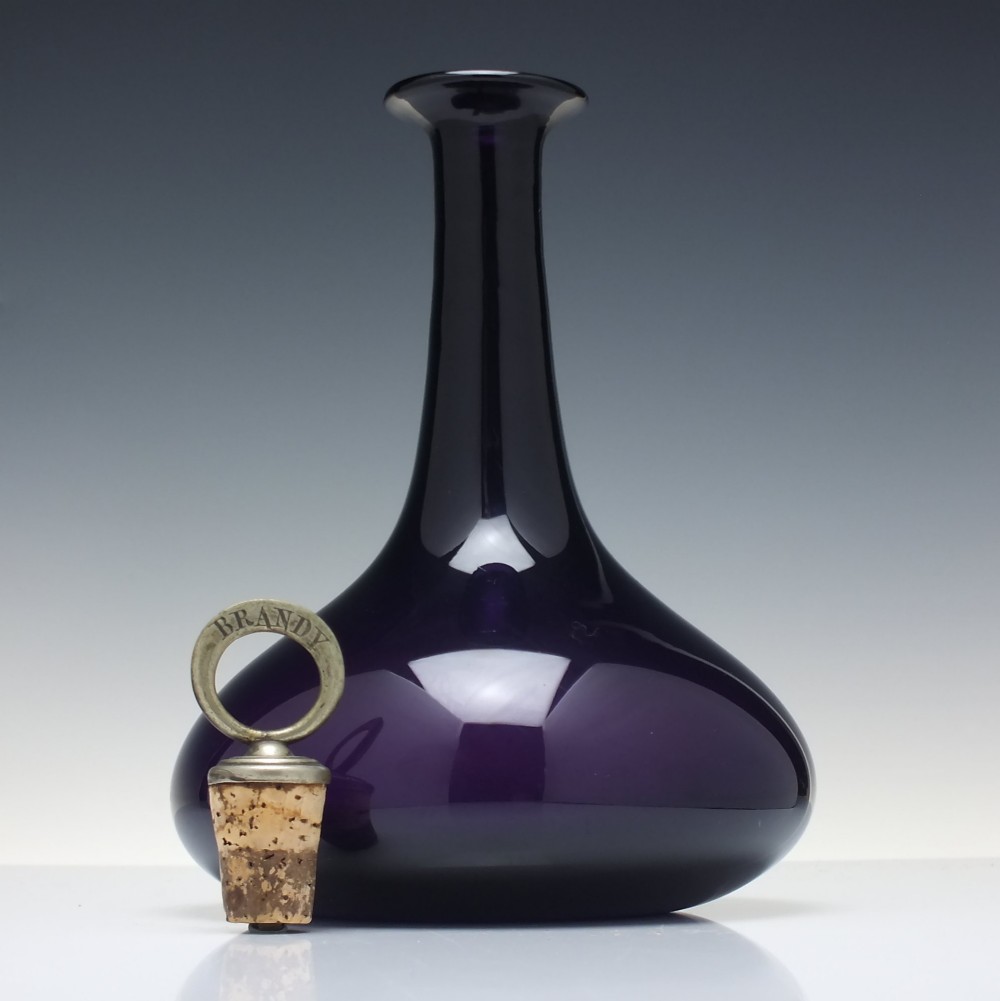 A deep purple amethyst glass decanter featuring a wide bulbous base, elongated neck, and a metal ring stopper engraved with the word BRANDY. View IV