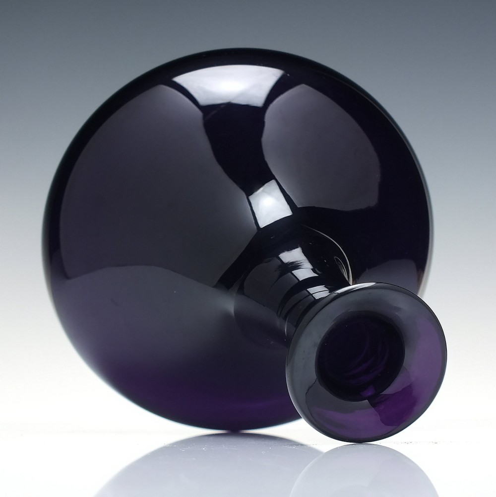 A deep purple amethyst glass decanter featuring a wide bulbous base, elongated neck, and a metal ring stopper engraved with the word BRANDY. View V