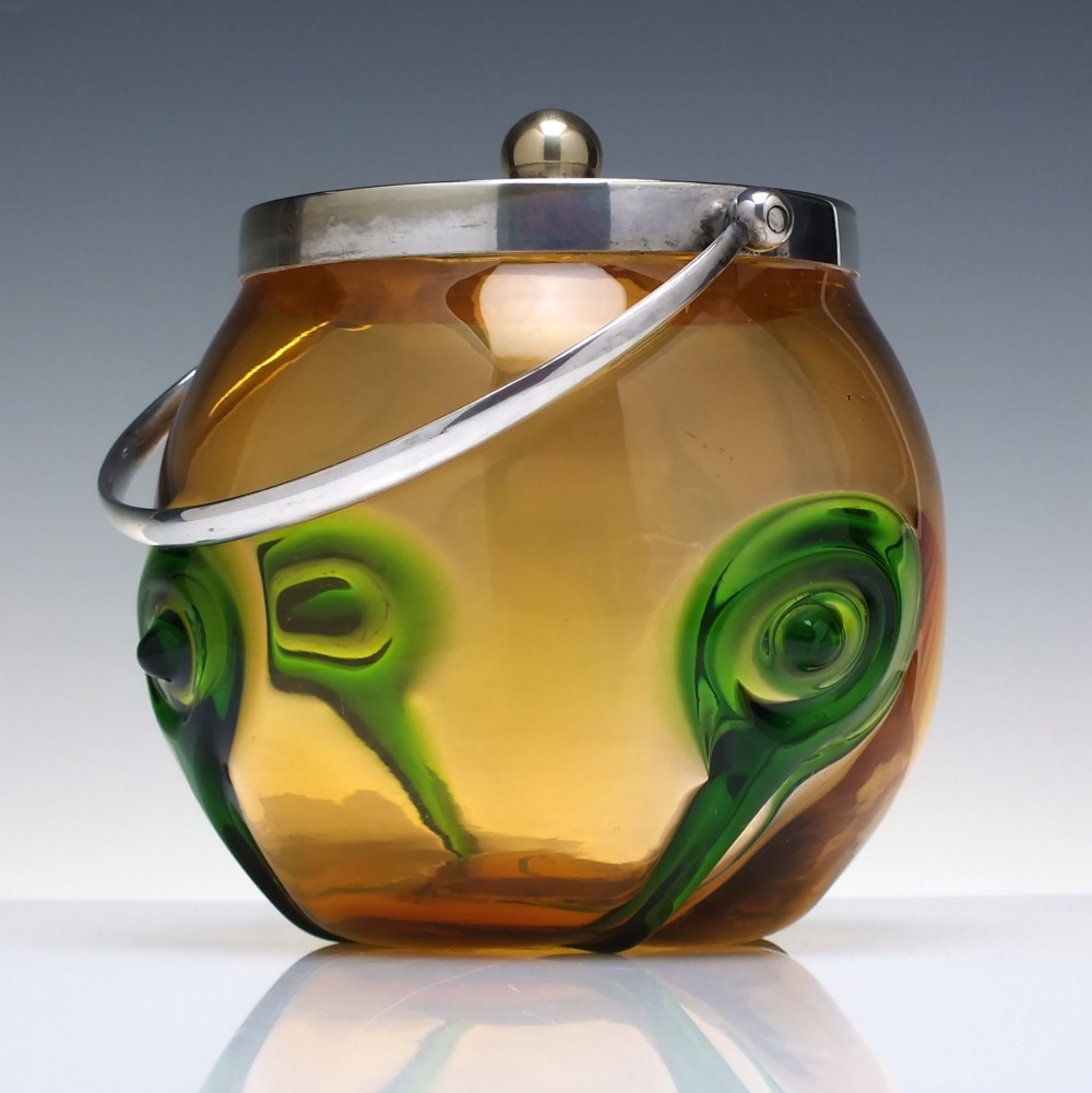 An Art Nouveau amber glass biscuit barrel featuring green peacock eye trailing decoration and a silver-plated metal rim, lid, and swing handle.