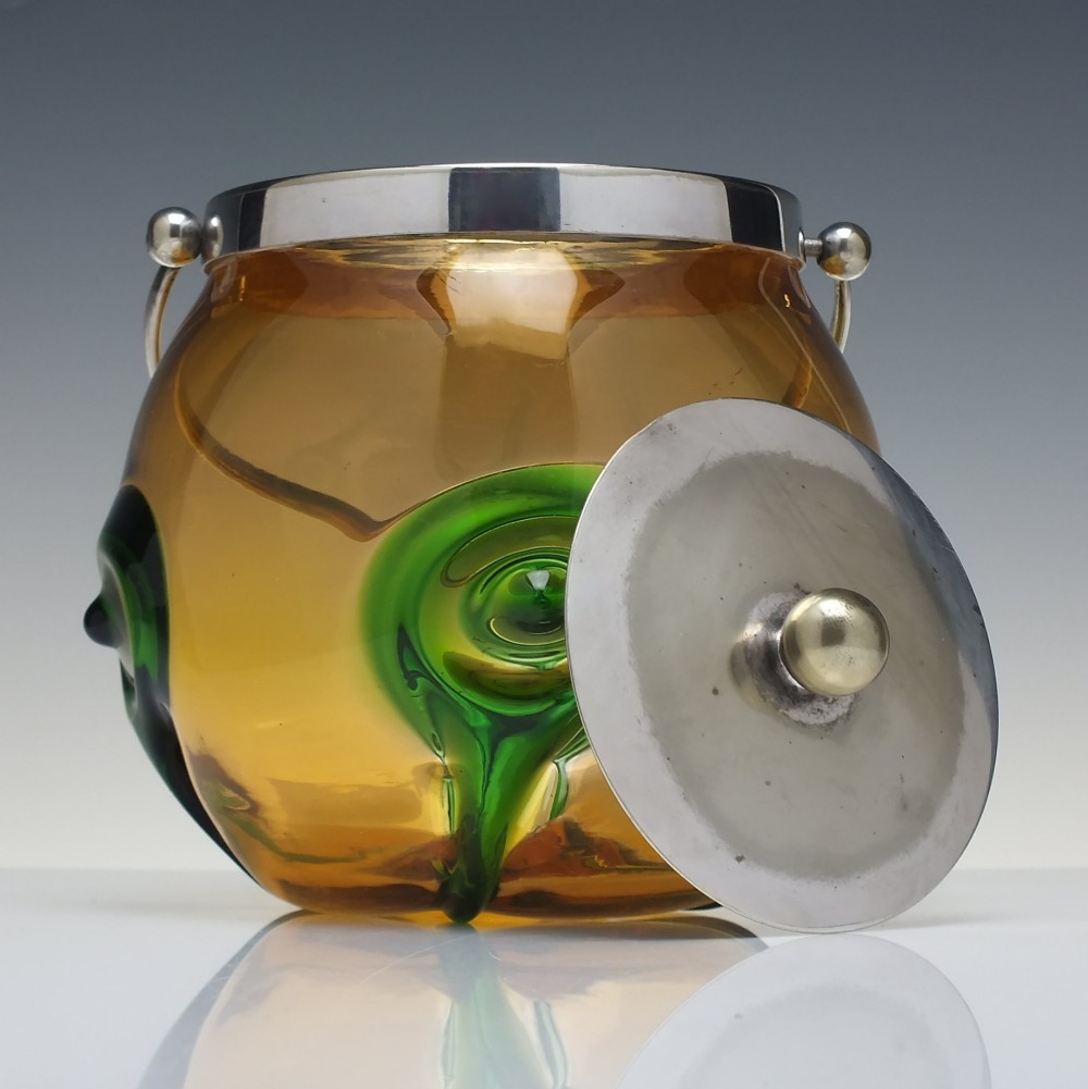 An Art Nouveau amber glass biscuit barrel featuring green peacock eye trailing decoration and a silver-plated metal rim, lid, and swing handle. View III