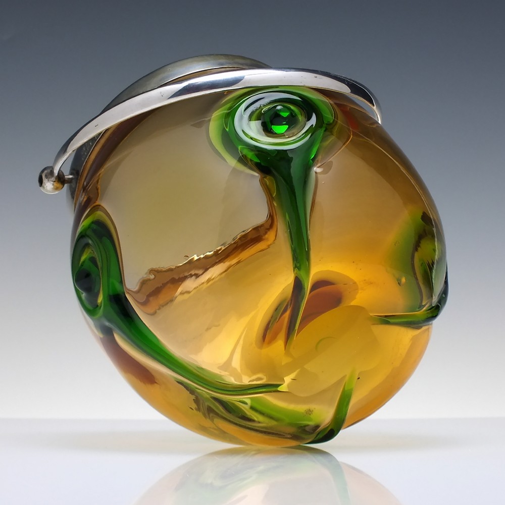 An Art Nouveau amber glass biscuit barrel featuring green peacock eye trailing decoration and a silver-plated metal rim, lid, and swing handle. View V
