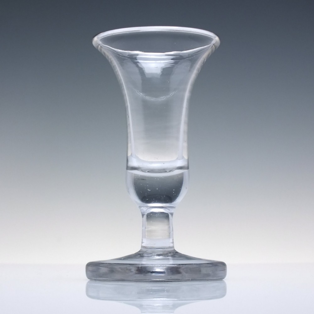 An 18th-century clear glass firing glass featuring a flared trumpet rim, short stem element, and a heavy, thick round foot. View II