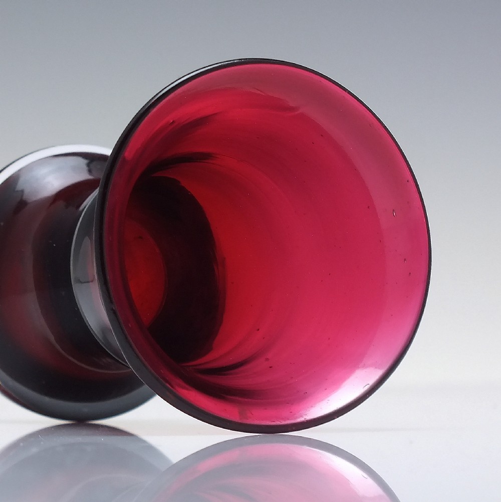 A small Victorian ruby glass dram tumbler with a flared lip and heavy thick base against a plain background. View III