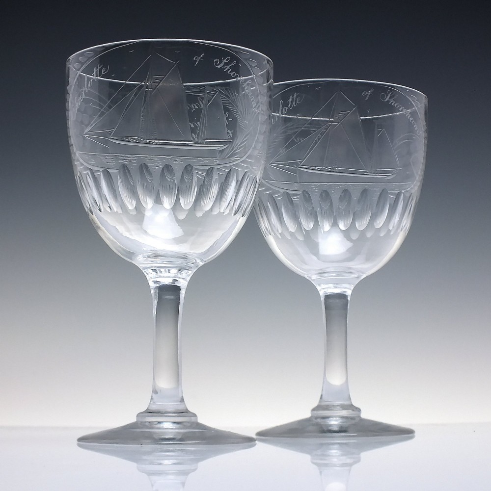 A pair of clear glass Victorian wine goblets featuring wheel-engraved sailing ships, inscriptions, and cut oval fluting around the lower bowls.