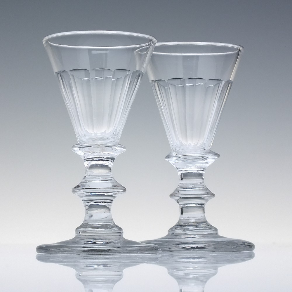 A pair of clear cut-glass Georgian dram glasses featuring conical bowls with vertical flutes and knopped stems on circular footed bases.