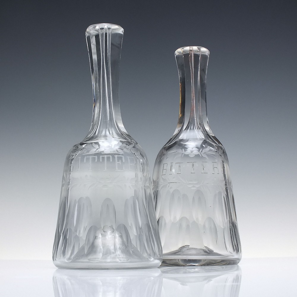 A pair of 18th-century clear glass sugar loaf bitters bottles featuring facet-cut long necks, hand-engraved floral garlands, and the word BITTERS cut into the body.