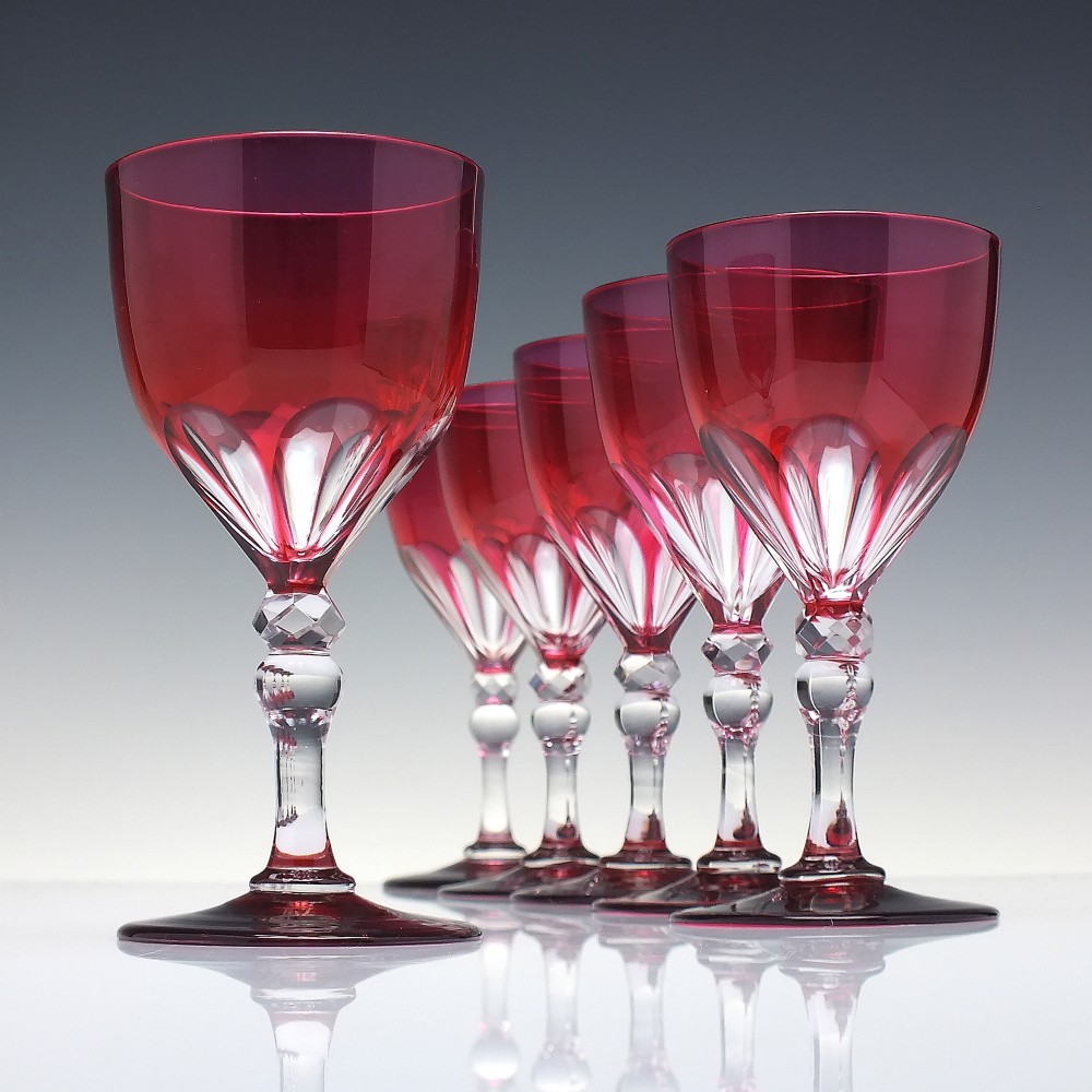 A line of six Victorian cranberry glass wine goblets featuring cut-to-clear patterned bases and faceted clear stems.
