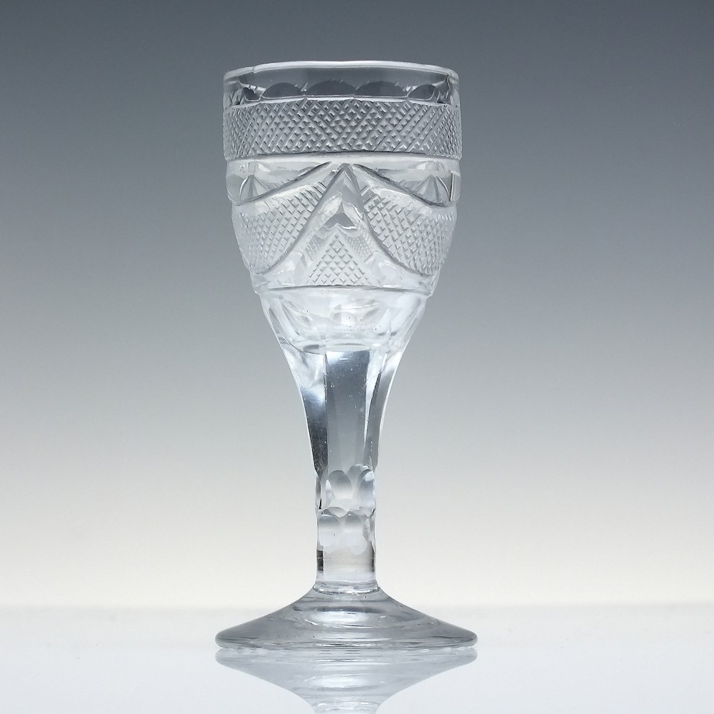 An antique clear cut lead crystal dram glass featuring diamond lattice cutting on the bowl, a faceted stem, and a flat circular base. View II
