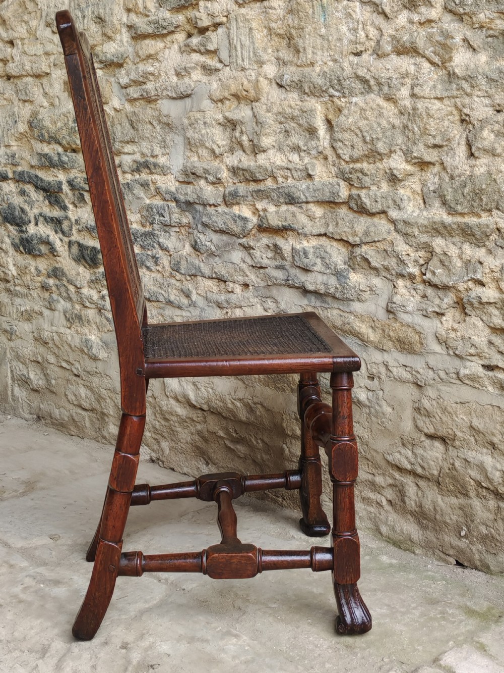 An early 18th-century walnut side chair featuring a woven cane backrest with a vertical central splat, a cane seat, turned legs, and a carved front stretcher. View VII