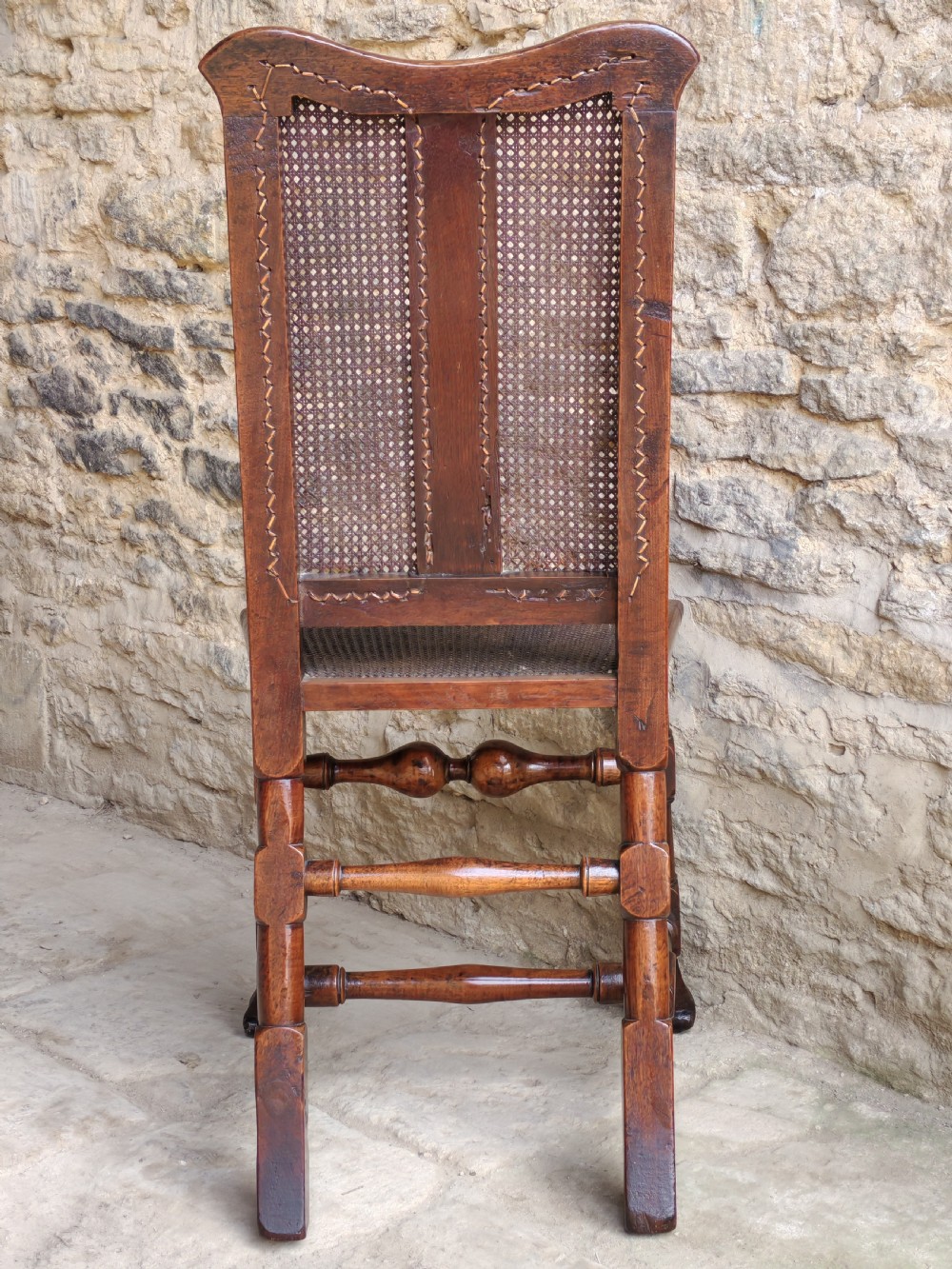An early 18th-century walnut side chair featuring a woven cane backrest with a vertical central splat, a cane seat, turned legs, and a carved front stretcher. View VIII