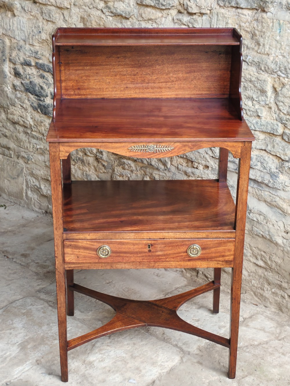 A Georgian mahogany bedside stand featuring a gallery back shelf, an open middle compartment, a single drawer with brass ring handles, and tapered legs joined by a cross-stretcher. View II