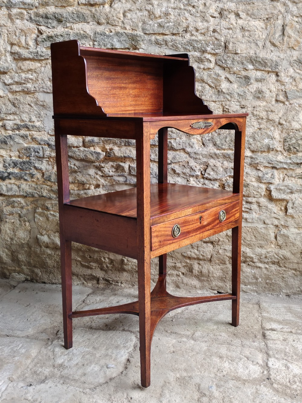 A Georgian mahogany bedside stand featuring a gallery back shelf, an open middle compartment, a single drawer with brass ring handles, and tapered legs joined by a cross-stretcher.