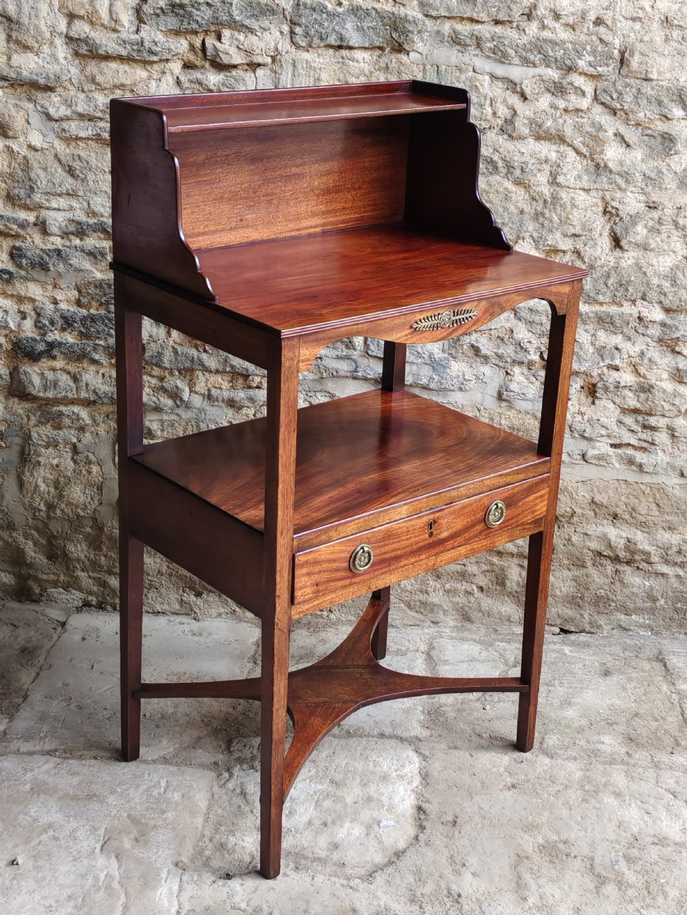 A Georgian mahogany bedside stand featuring a gallery back shelf, an open middle compartment, a single drawer with brass ring handles, and tapered legs joined by a cross-stretcher. View VII