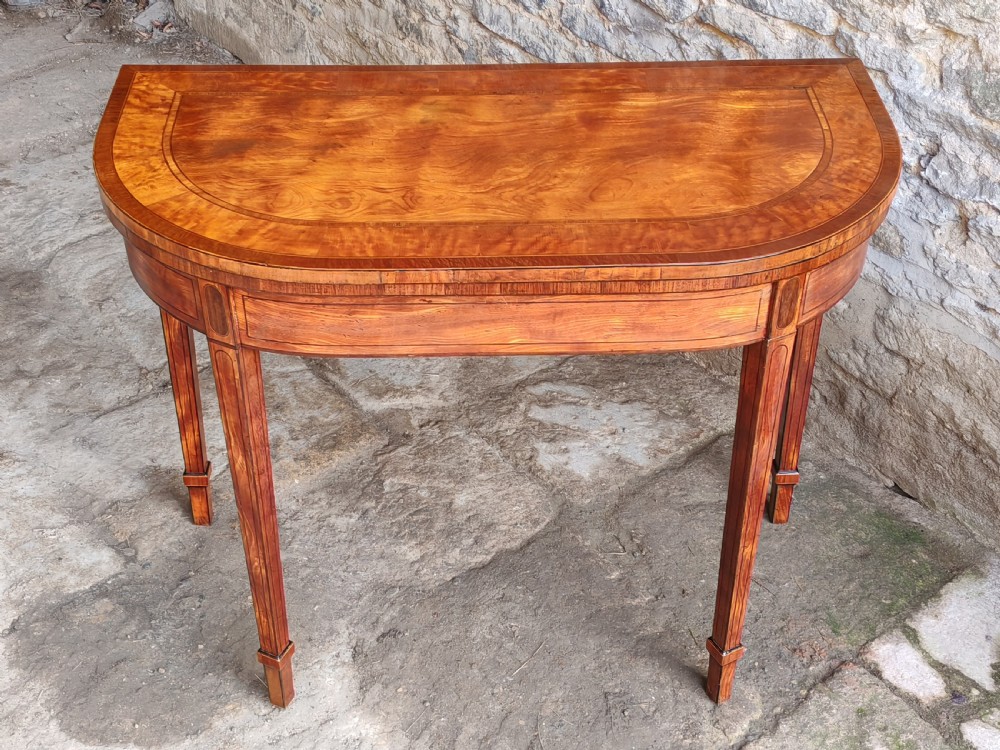 A late eighteenth-century Georgian satinwood demi-lune card table featuring inlaid crossbanding and four square tapered legs.