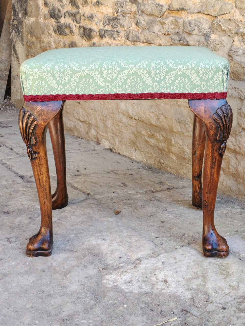 A rectangular 19th-century walnut stool featuring cabriole legs with carved shell details and a light green patterned fabric seat edged with red braid trim. View II