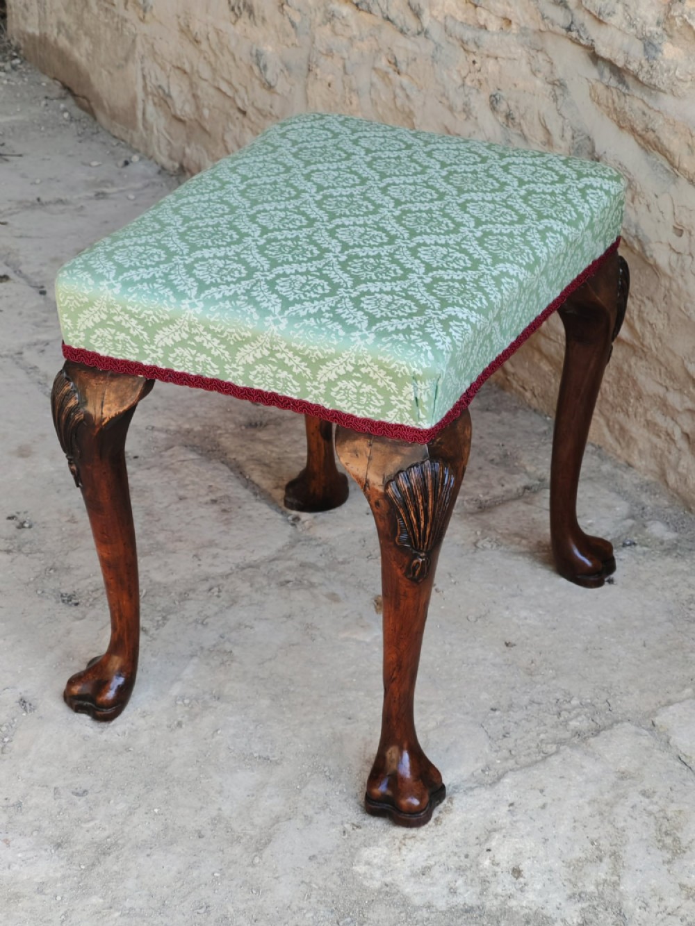 A rectangular 19th-century walnut stool featuring cabriole legs with carved shell details and a light green patterned fabric seat edged with red braid trim. View VII