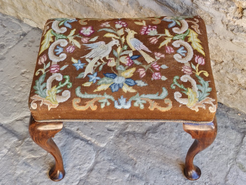 An antique walnut footstool with cabriole legs featuring a brown needlepoint upholstered seat depicting two birds amidst flowers.