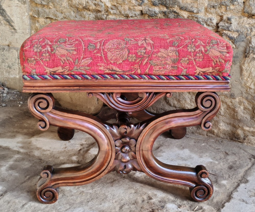 A Victorian mahogany X-frame stool featuring carved scrollwork legs, a central floral rosette motif, and a padded red floral upholstered seat with corded trim.