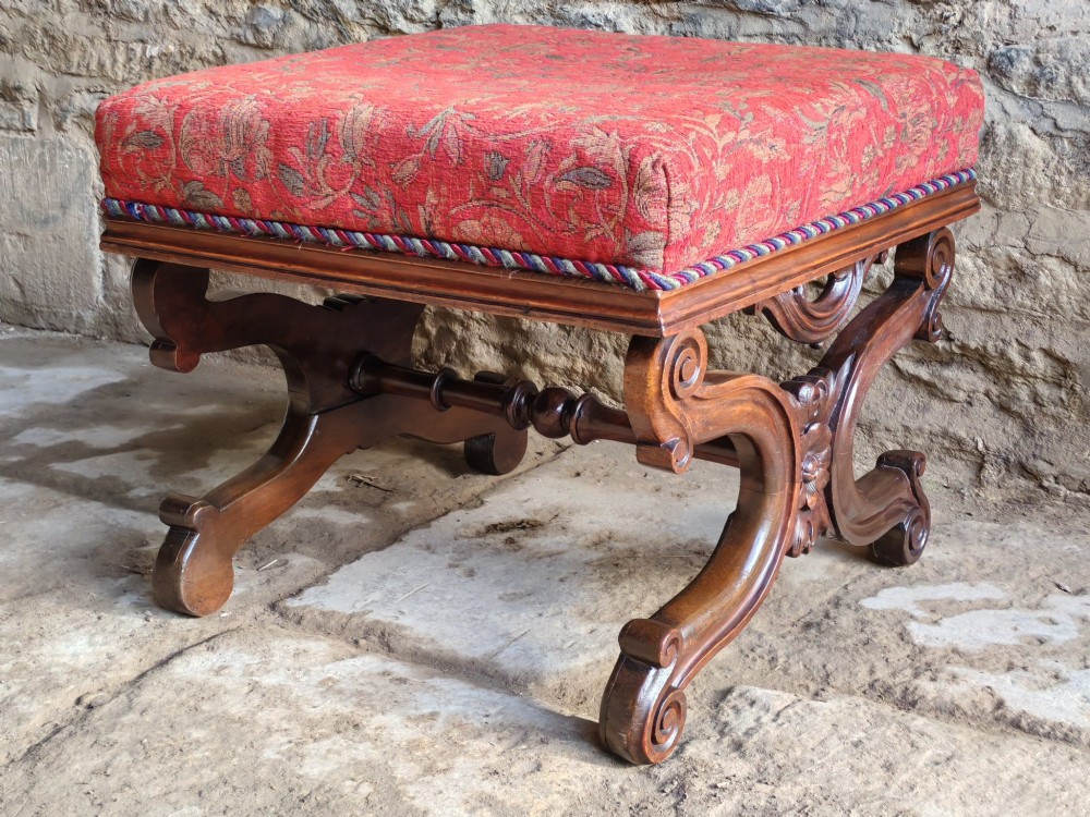 A Victorian mahogany X-frame stool featuring carved scrollwork legs, a central floral rosette motif, and a padded red floral upholstered seat with corded trim. View II