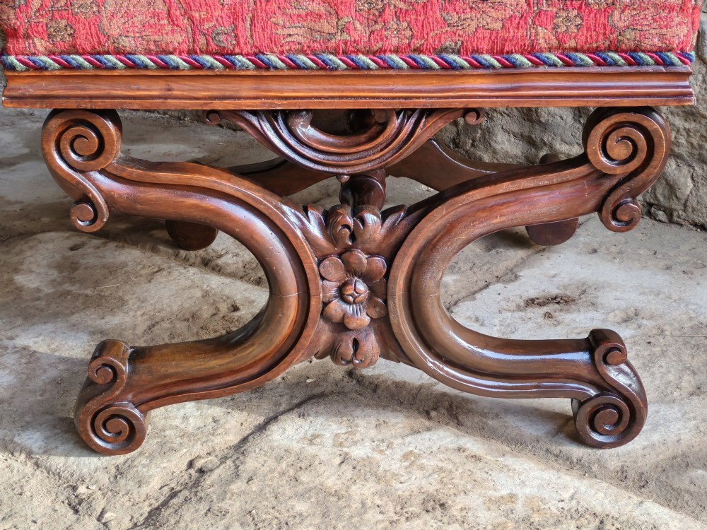 A Victorian mahogany X-frame stool featuring carved scrollwork legs, a central floral rosette motif, and a padded red floral upholstered seat with corded trim. View III