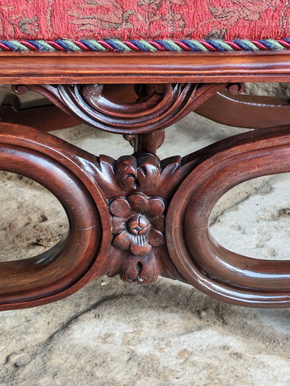 A Victorian mahogany X-frame stool featuring carved scrollwork legs, a central floral rosette motif, and a padded red floral upholstered seat with corded trim. View IV