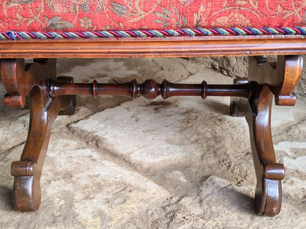 A Victorian mahogany X-frame stool featuring carved scrollwork legs, a central floral rosette motif, and a padded red floral upholstered seat with corded trim. View V