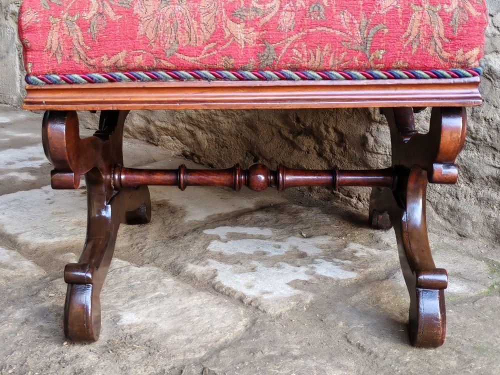 A Victorian mahogany X-frame stool featuring carved scrollwork legs, a central floral rosette motif, and a padded red floral upholstered seat with corded trim. View VI