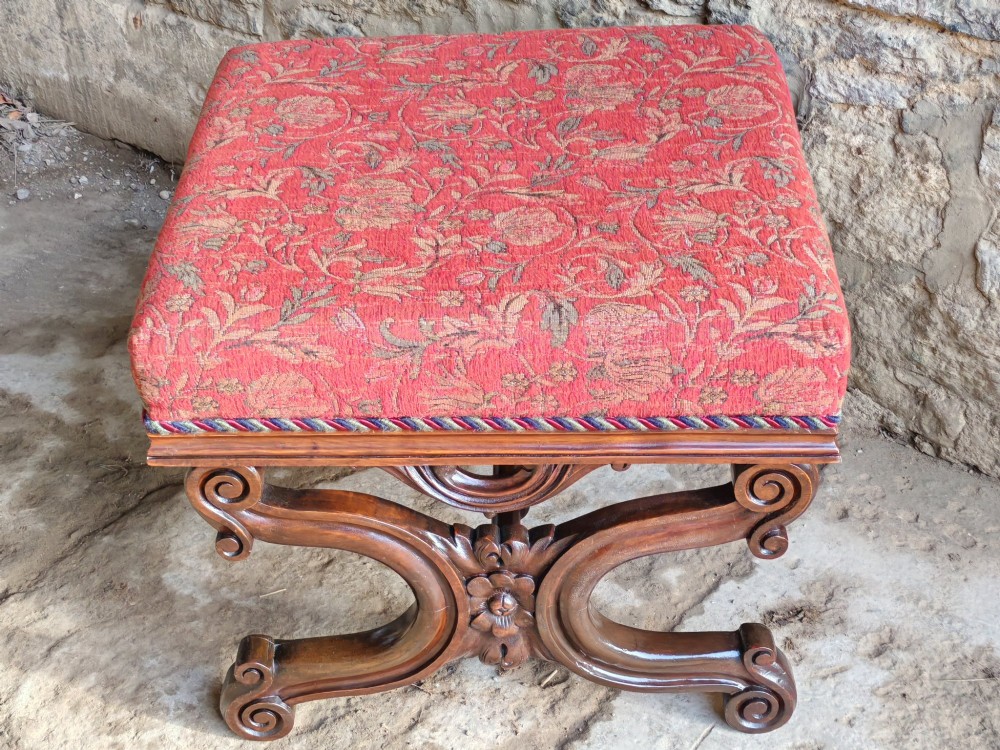 A Victorian mahogany X-frame stool featuring carved scrollwork legs, a central floral rosette motif, and a padded red floral upholstered seat with corded trim. View VII