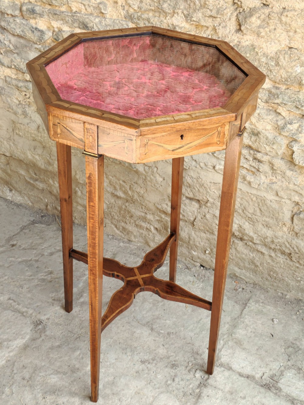 An octagonal late Victorian wood bijouterie table with marquetry swags, a glazed top displaying a pink fabric lining, tapering square legs, and a shaped cross-stretcher.