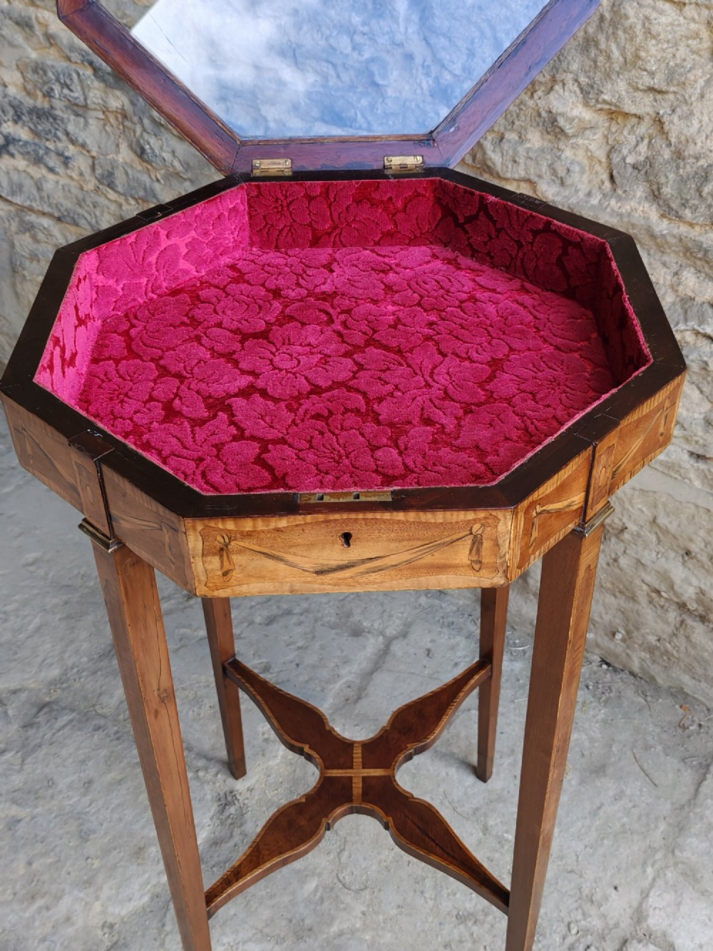 An octagonal late Victorian wood bijouterie table with marquetry swags, a glazed top displaying a pink fabric lining, tapering square legs, and a shaped cross-stretcher. View V