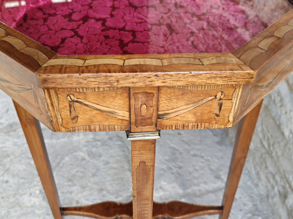 An octagonal late Victorian wood bijouterie table with marquetry swags, a glazed top displaying a pink fabric lining, tapering square legs, and a shaped cross-stretcher. View VIII