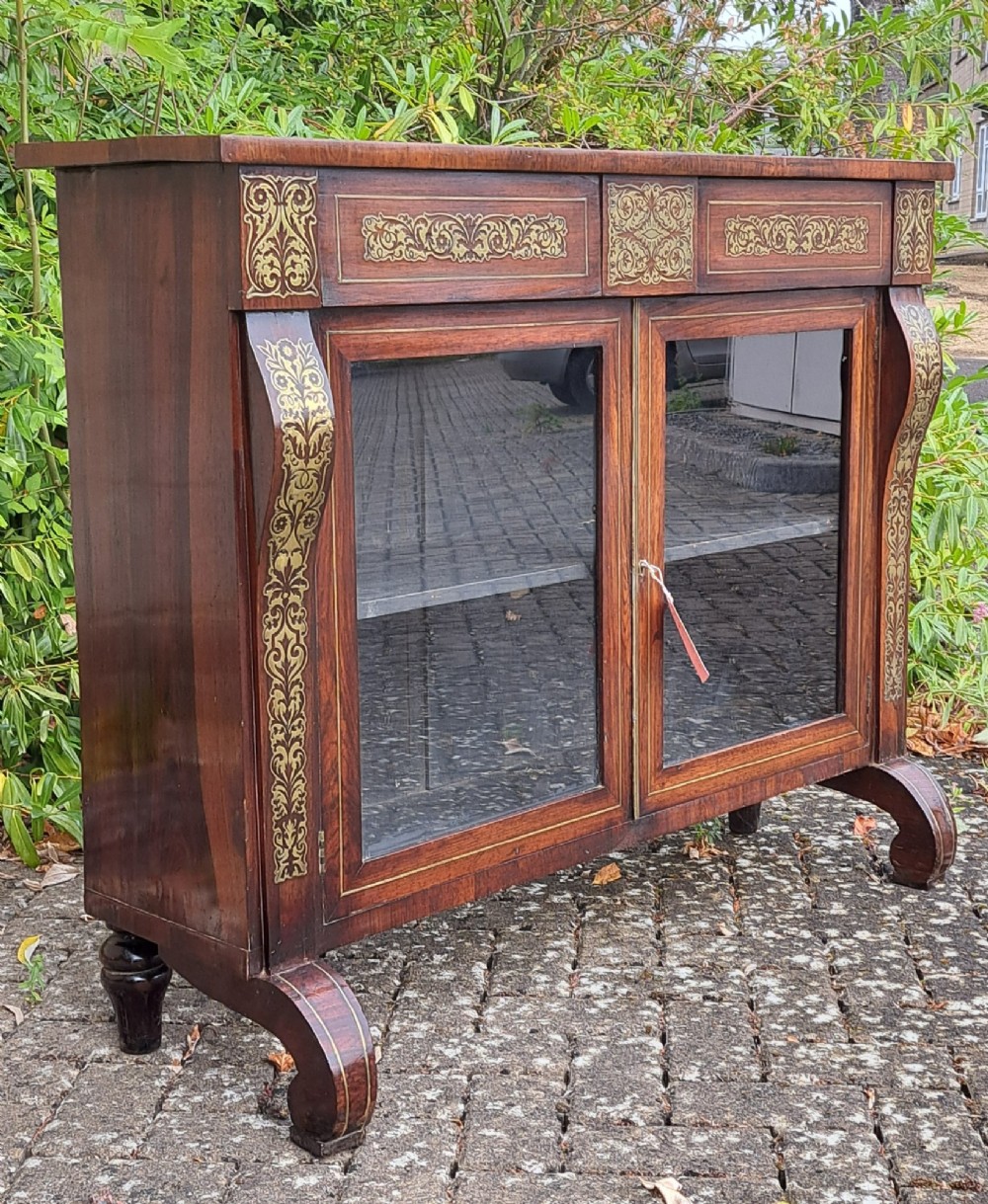 An 1830s rosewood low bookcase featuring twin glazed doors, scroll feet, and foliate brass inlay across the frieze and pilasters.