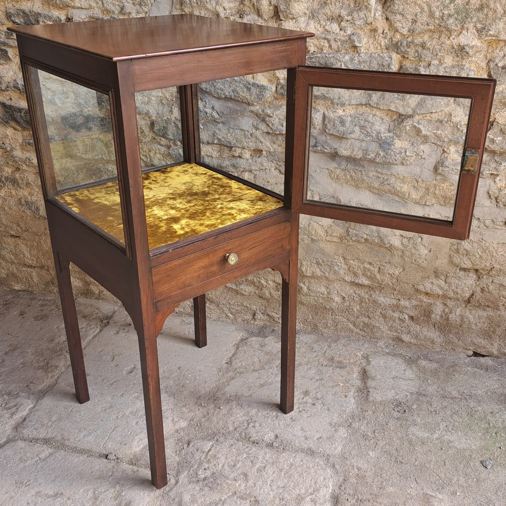 A Georgian mahogany standing display cabinet featuring a glass-enclosed upper section lined with gold fabric, a single lower drawer, and square tapered legs.